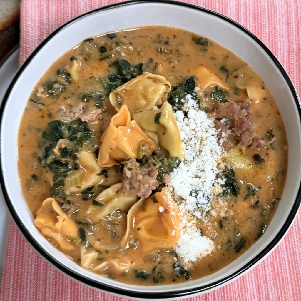 Creamy Sausage and Tortellini Soup with Spinach with grated cheese in soup bowl over red towel.