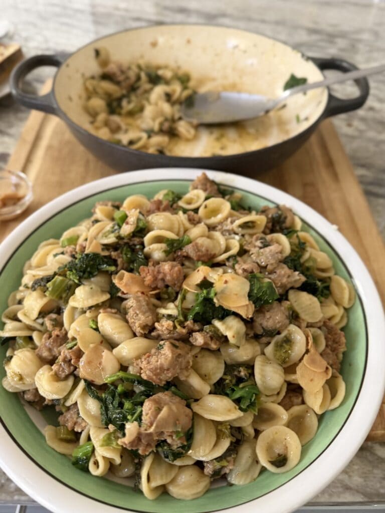 Rapini with Sausage and Pasta in large pasta serving bowl with pan and spoon in the background.