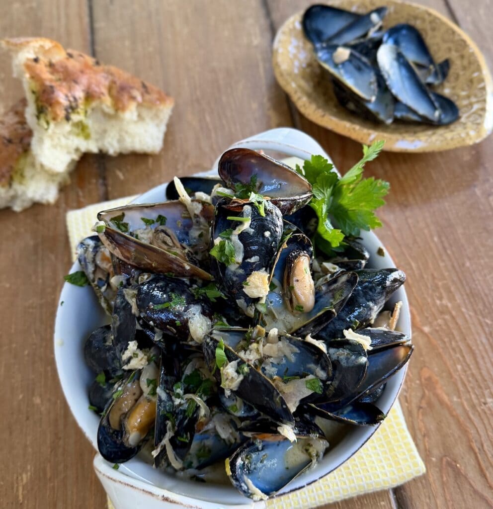 French style mussels (moules mariniere) in white serving dish with parsley garnish and dish with empty mussel shells and focaccia bread in background.