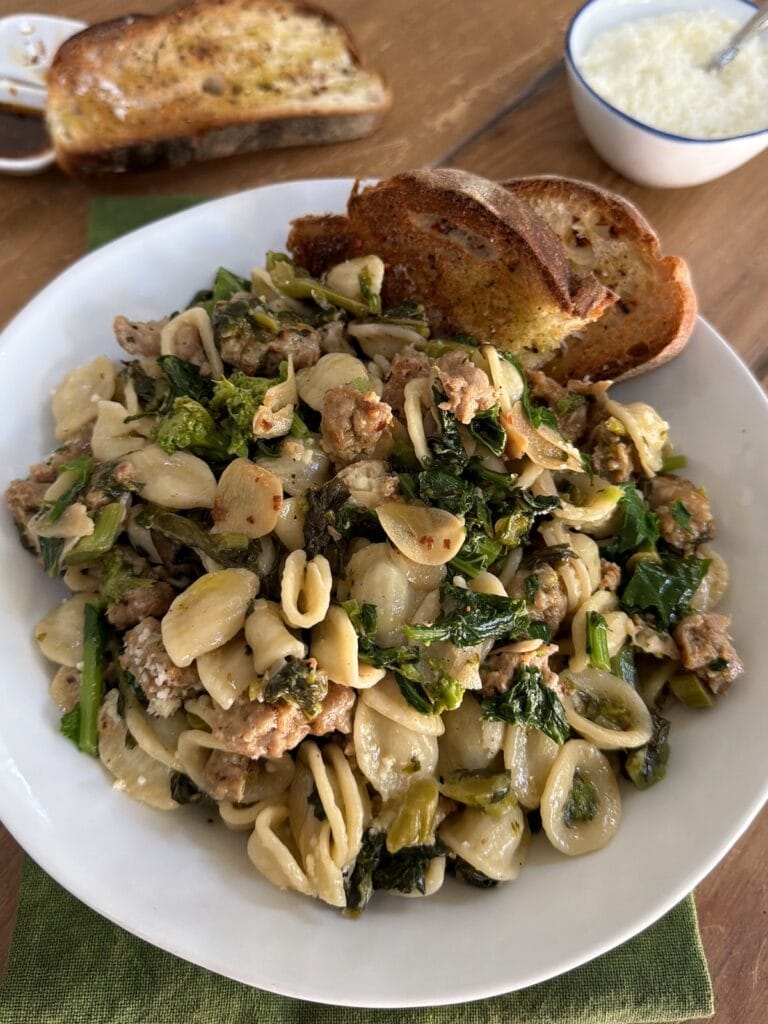Rapini Sausage Pasta in white bowl with two slices of bread with small bowl of grated cheese and spicy oil and bread in background.