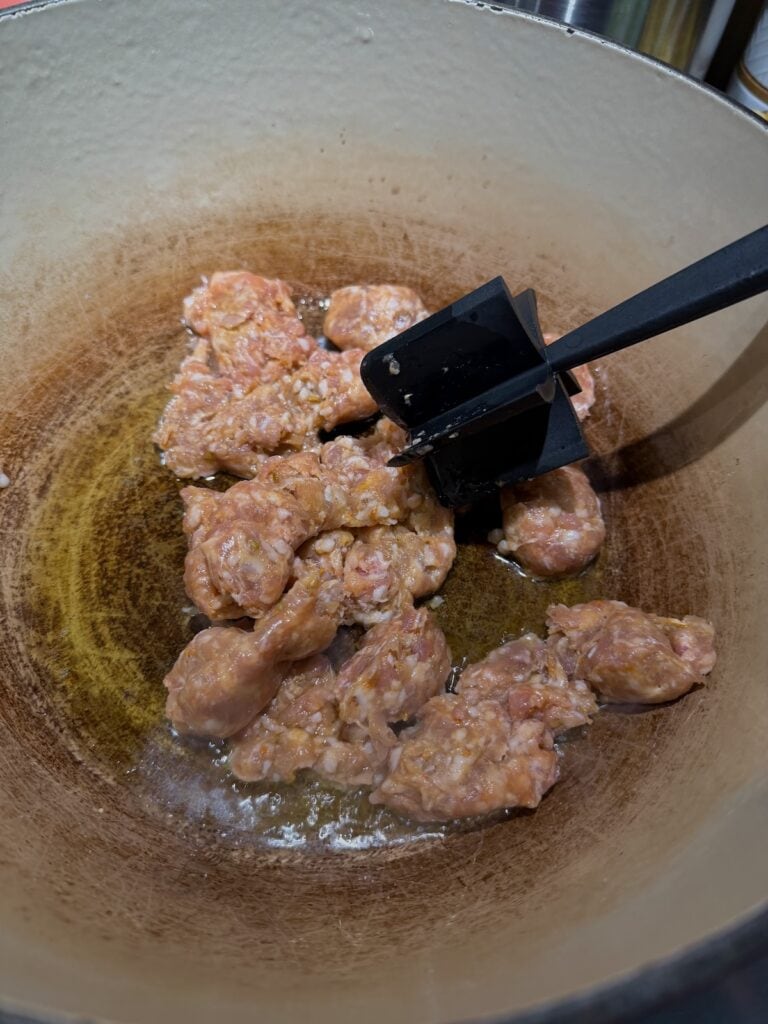 Browning sausage in Dutch oven and breaking it up into smaller pieces using a meat masher.