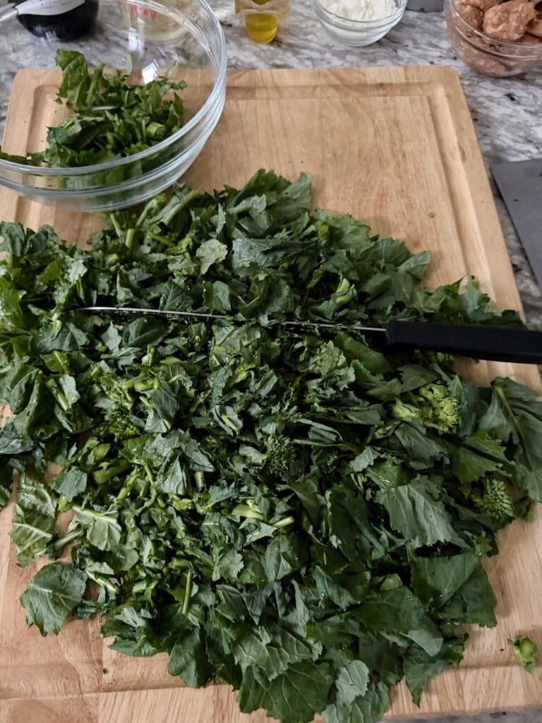 Chopping broccoli di rape leaves and flowers on cutting board with knife into bite-sized pieces.