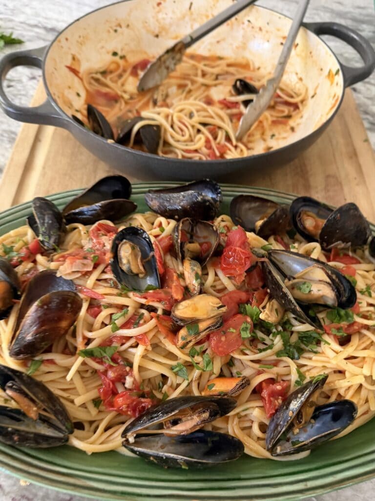 Italian American mussels and linguine in green serving platter with pan and tongs in the background.