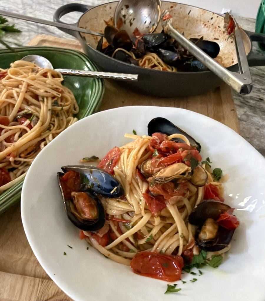 Individual bowl of mussels pasta with platter and pan full of pasta in background.