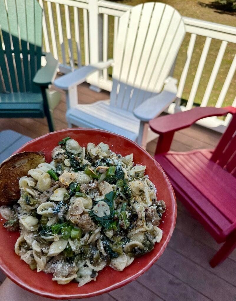 Orecchiette With Broccoli Rabe and Sausage in orange bowl with bread with deck chairs in the background.