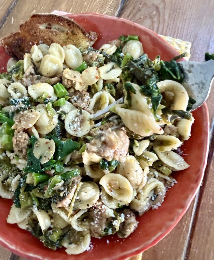 Rapini with Sausage and Pasta in orange bowl with slice of bread, with forkful of mixture over it.