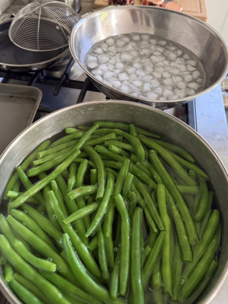 Pot of string beans in boiling water on stove next to bowl of ice water.