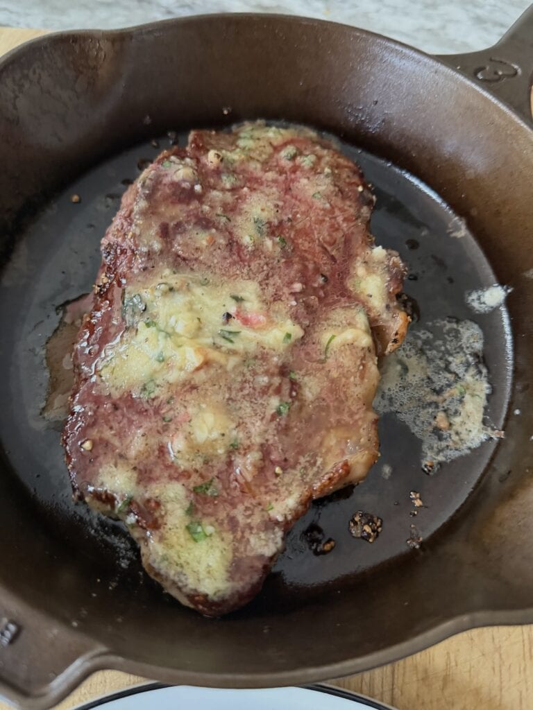 Garlic herb butter melting over seared rib eye steak in cast iron pan.