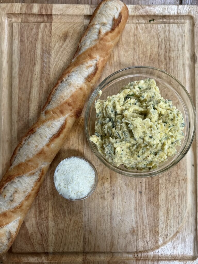 Overhead view of one baguette, and bowls of cheese and garlic butter on cutting board.