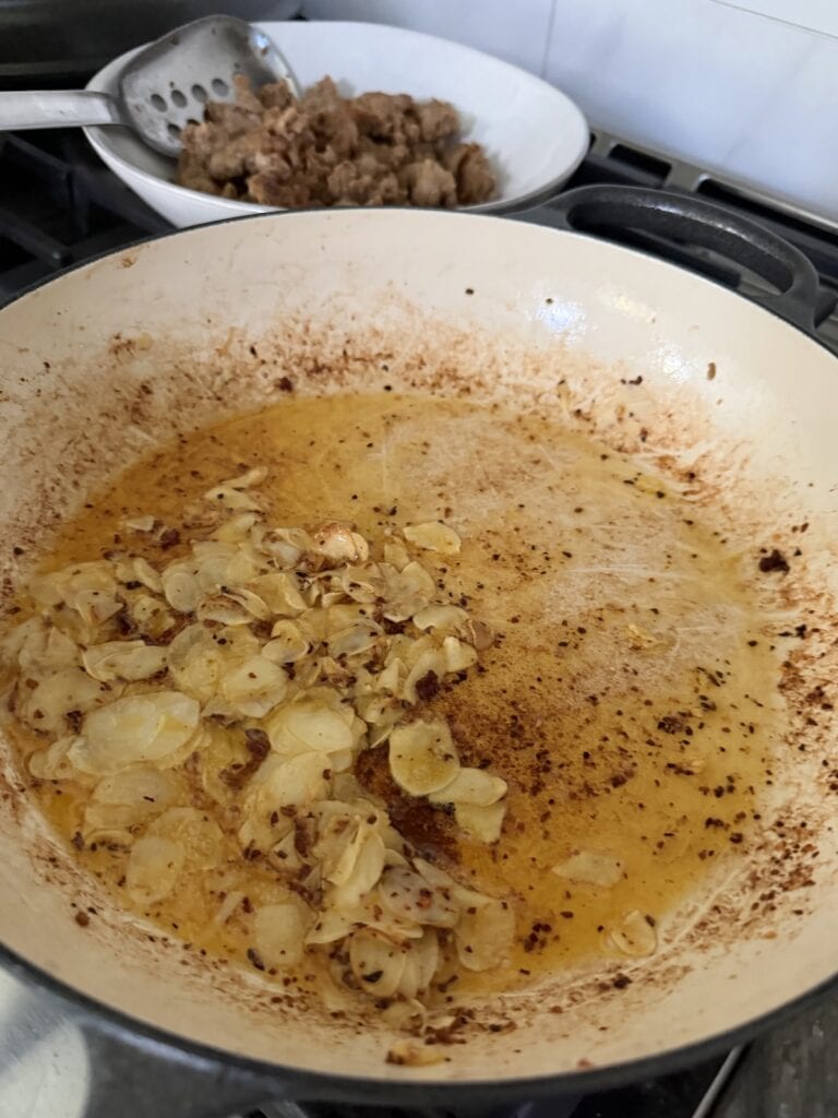 Sautéing thinly-sliced grlic in oil and sausage drippings in pan.