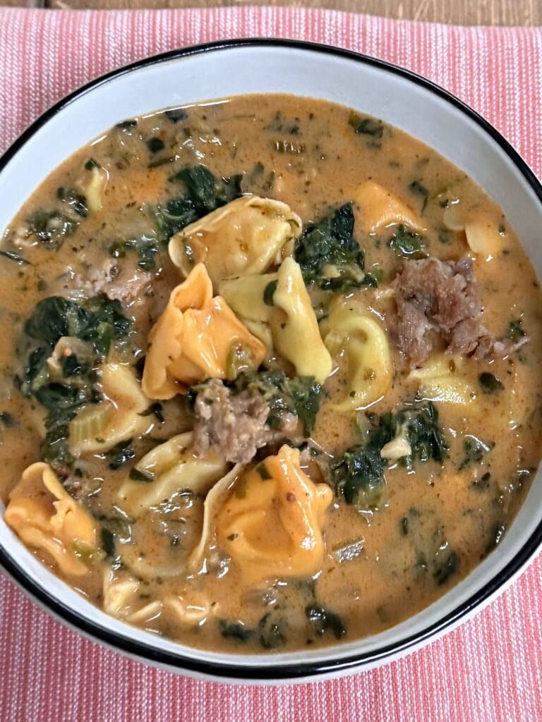 Bowl of creamy Italian sausage tortellini soup with spinach and cheese tortellini over red towel.