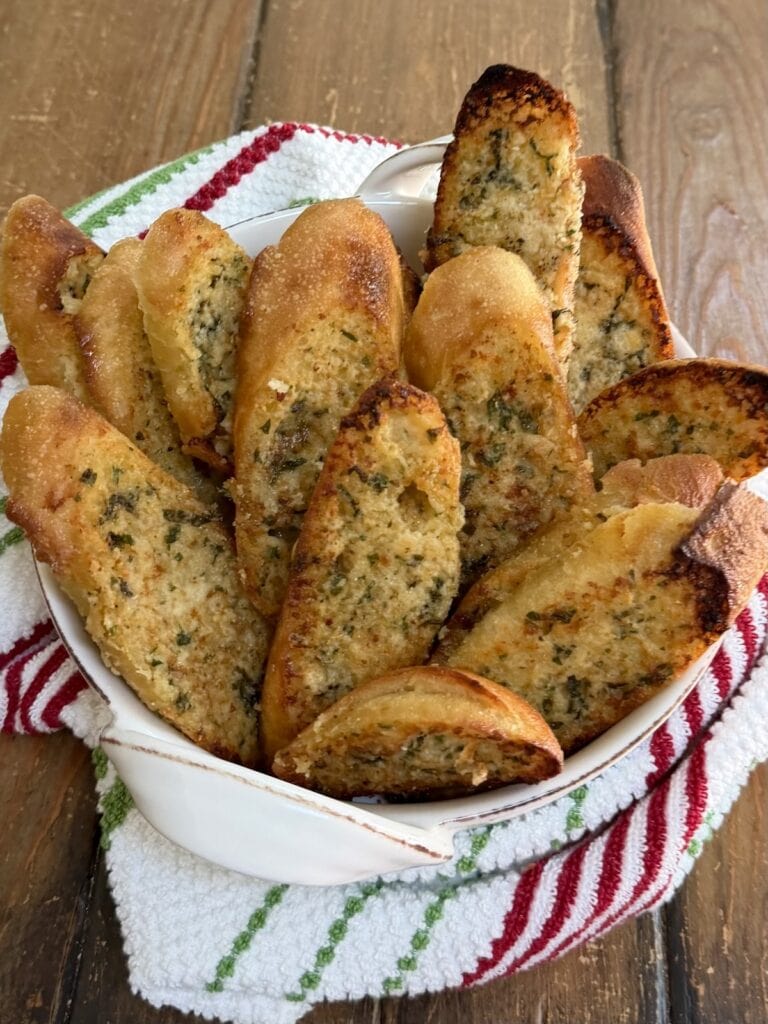Several slices of homemade garlic bread baguette in serving bowl with decorative towel.